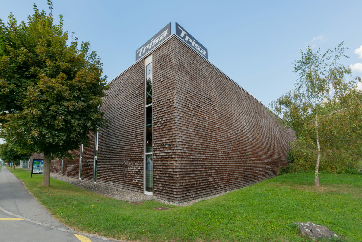 Betriebsgebäude von Trisa Accessoires AG in Triengen Schweiz, ein Holzschindelgebäude mit Photovoltaik für mehr Nachhaltigkeit, bei blauem Himmel und mit grüner Wiese